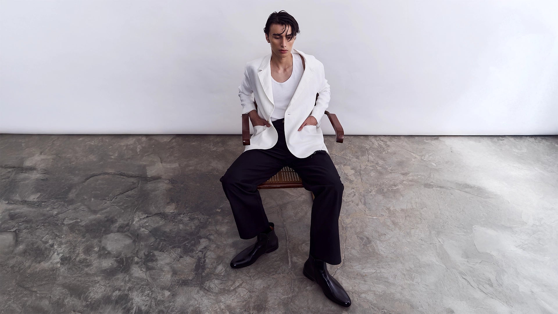 Person sitting on a chair wearing a white shirt and black pants in a minimalistic setting.