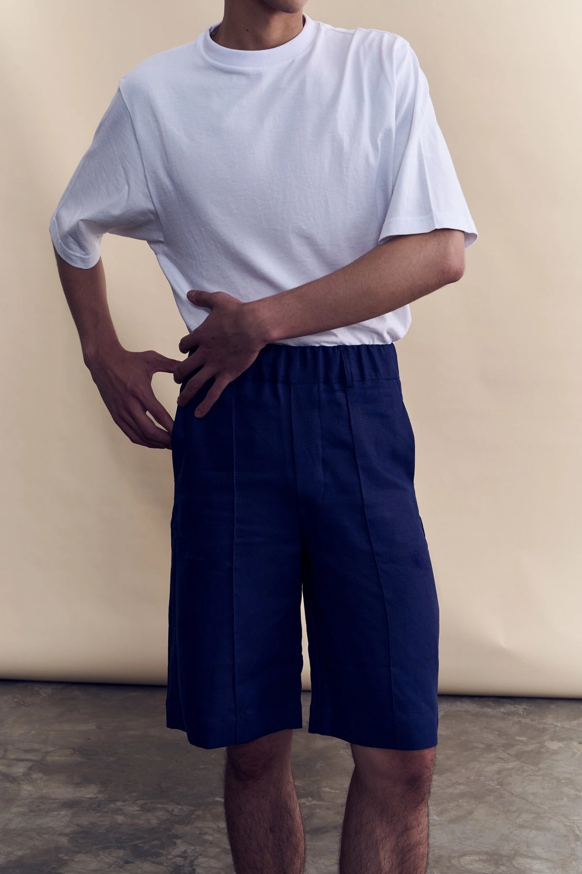 A model wearing a white short-sleeve crewneck t-shirt and navy blue shorts, standing against a beige studio backdrop.