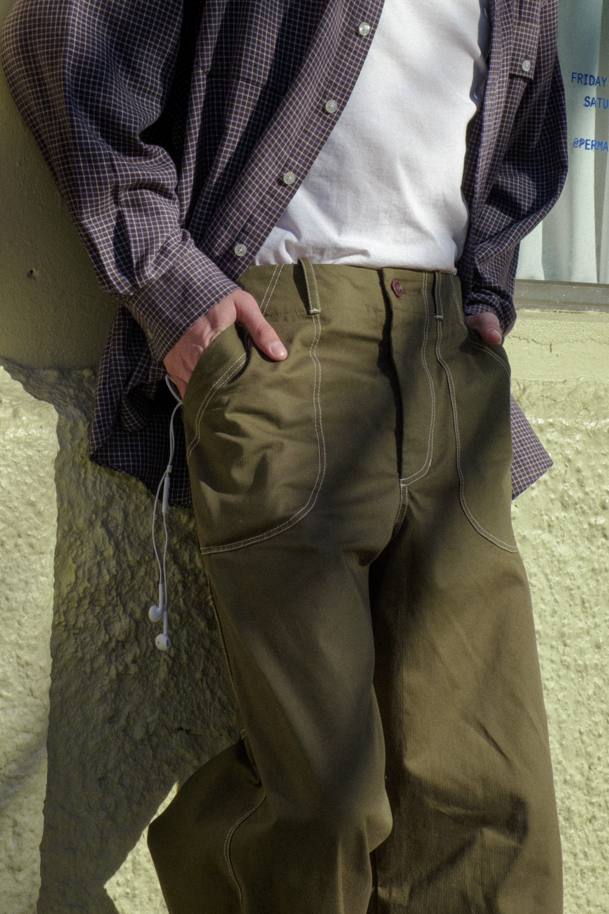 A man wearing olive green fatigue trousers with contrast stitching, a white t-shirt, and a dark checkered button-up shirt, leaning against a textured beige wall.