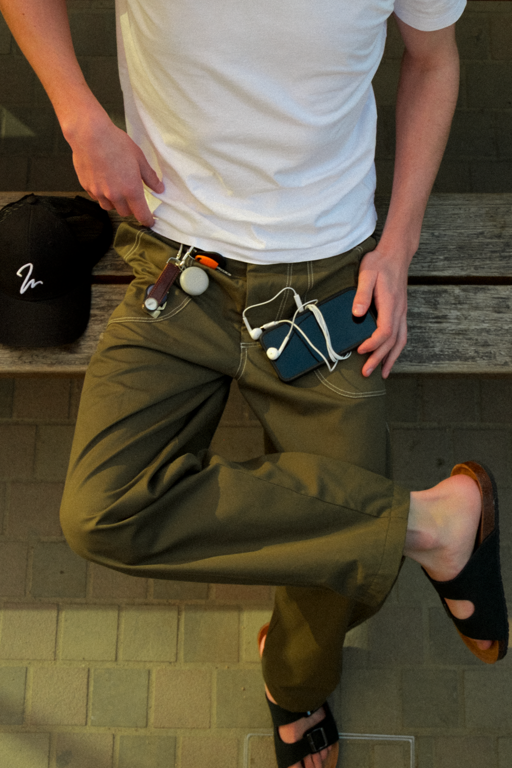 A man wearing olive green fatigue trousers with multiple accessories attached to the waistband, including a black cap, paired with a white t-shirt and sandals.