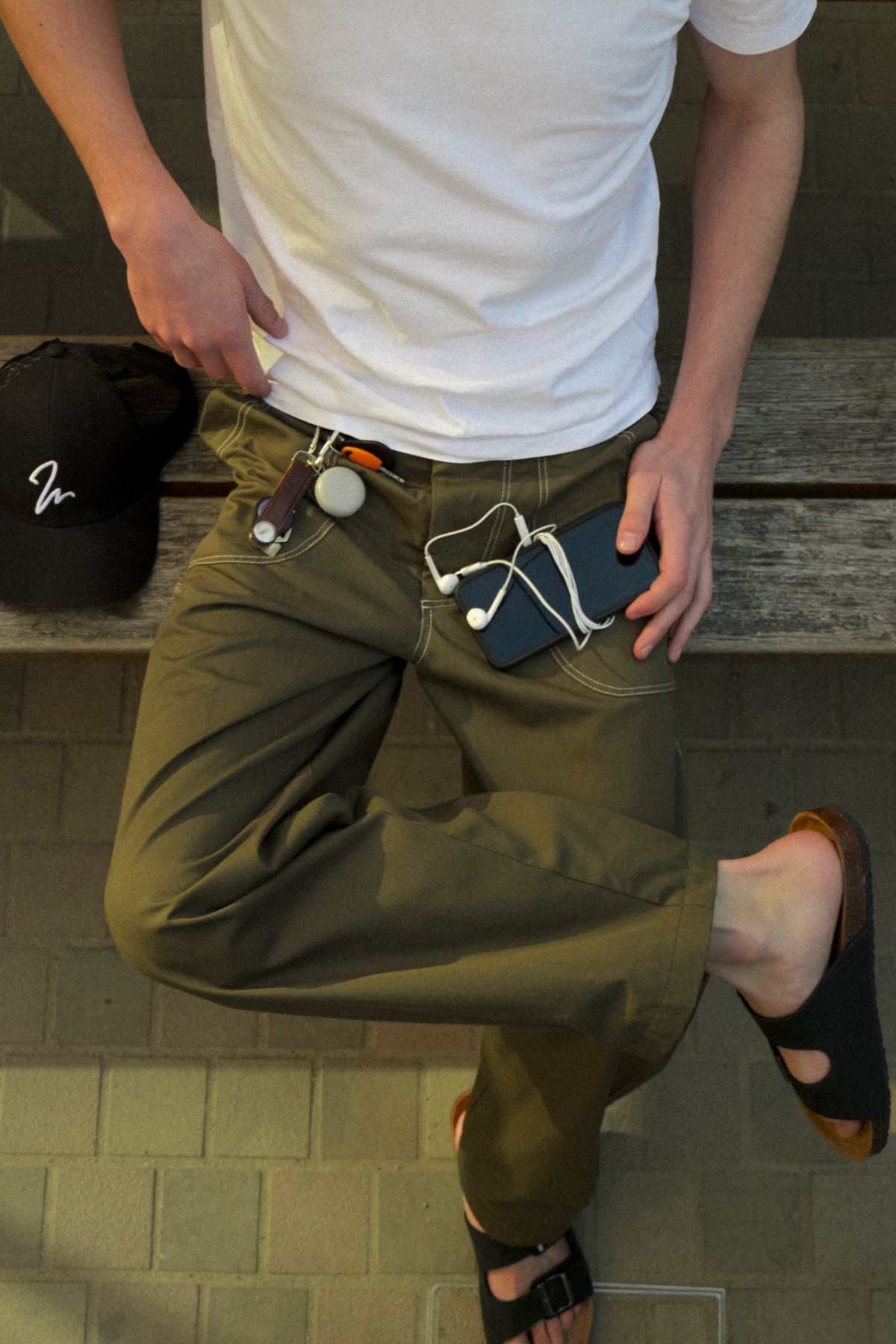 A man wearing olive green fatigue trousers with multiple accessories attached to the waistband, including a black cap, paired with a white t-shirt and sandals.