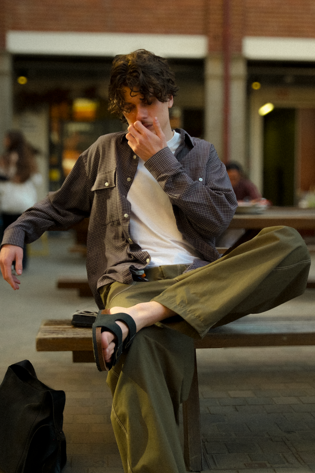 A man sits on a wooden bench in an outdoor urban setting, wearing a dark brown textured overshirt over a white t-shirt, olive green fatigue trousers, and  sandals.