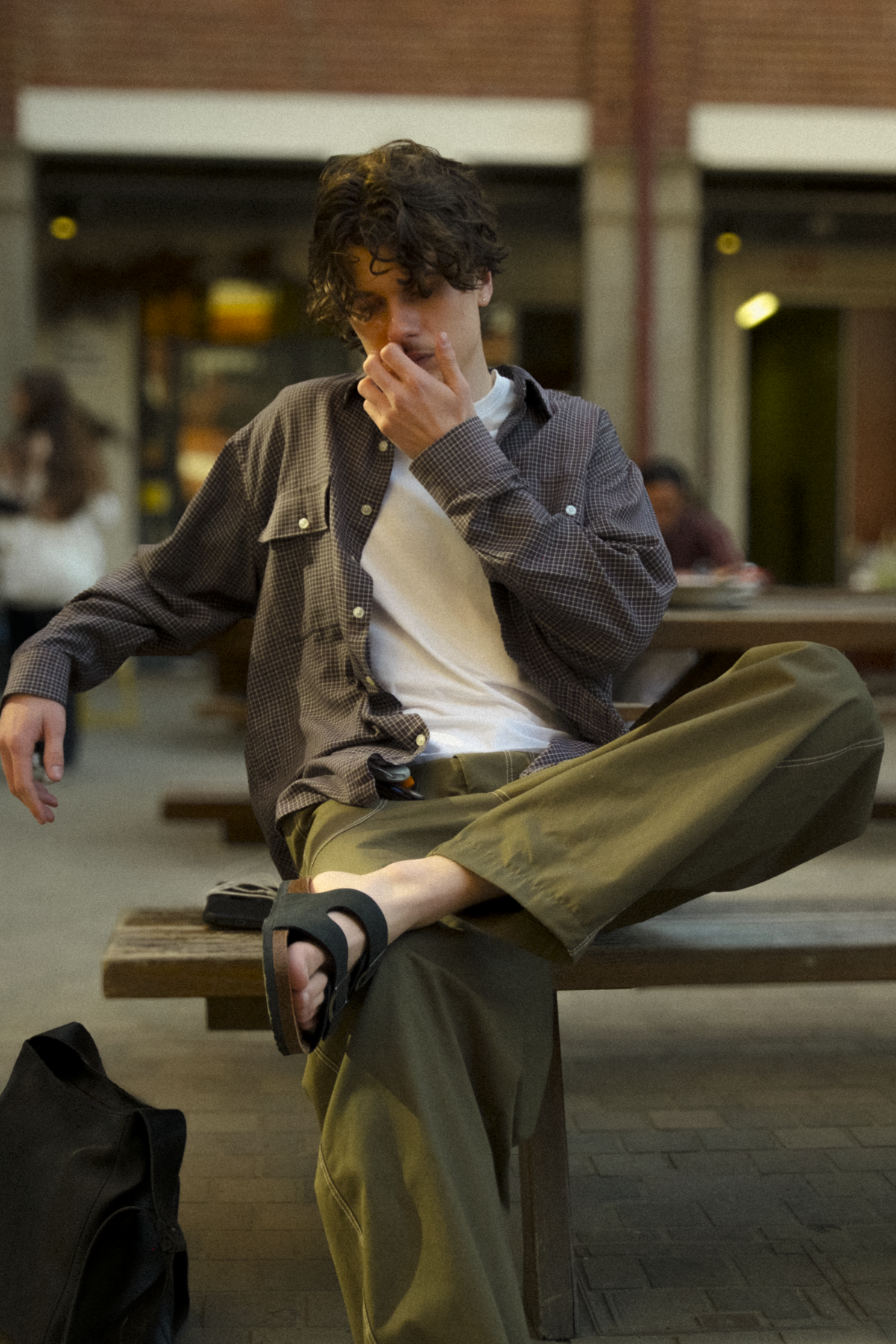 A man sits on a wooden bench in an outdoor urban setting, wearing a dark brown textured overshirt over a white t-shirt, olive green fatigue trousers, and  sandals.