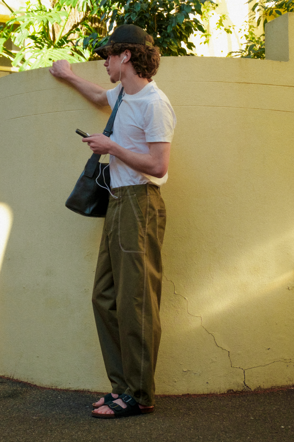A man wearing a white t-shirt, olive green fatigue trousers, a black trucker cap, and sandals stands against a beige stucco wall, holding a black shoulder bag and looking at his phone.