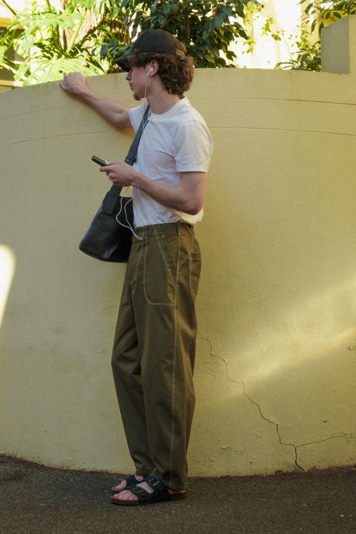 A man wearing a white t-shirt, olive green fatigue trousers, a black trucker cap, and sandals stands against a beige stucco wall, holding a black shoulder bag and looking at his phone.