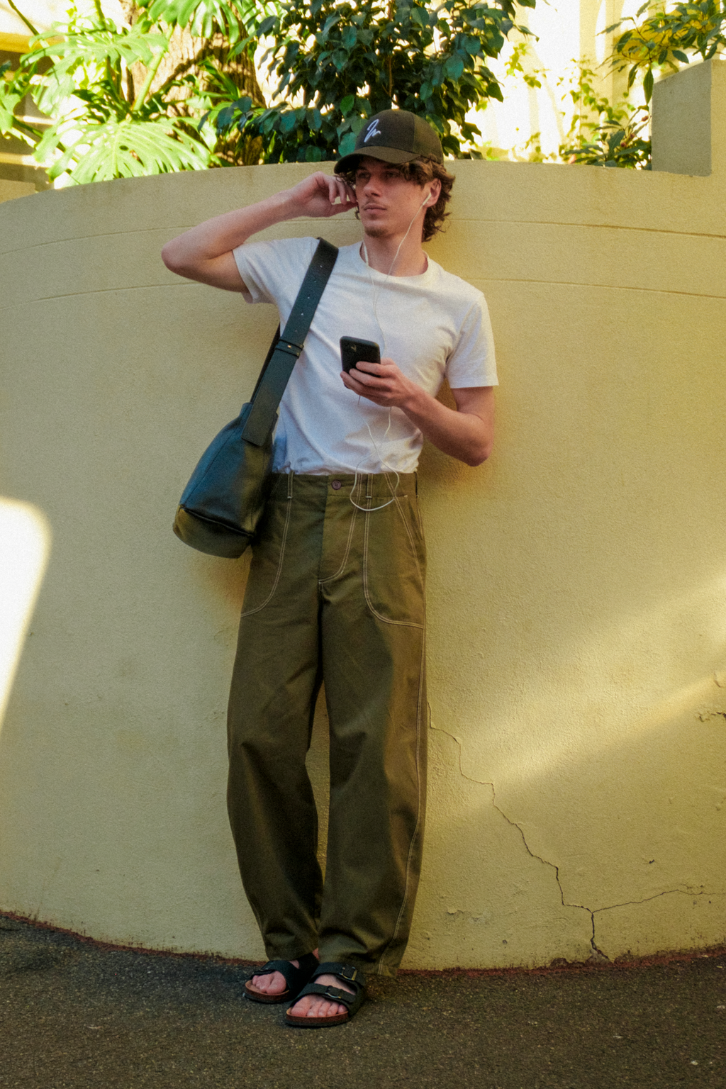 A man stands against a beige stucco wall outdoors, wearing a white t-shirt, olive green fatigue trousers, a black trucker cap, and sandals. He has a black shoulder bag slung over his shoulder and is holding a black smartphone.