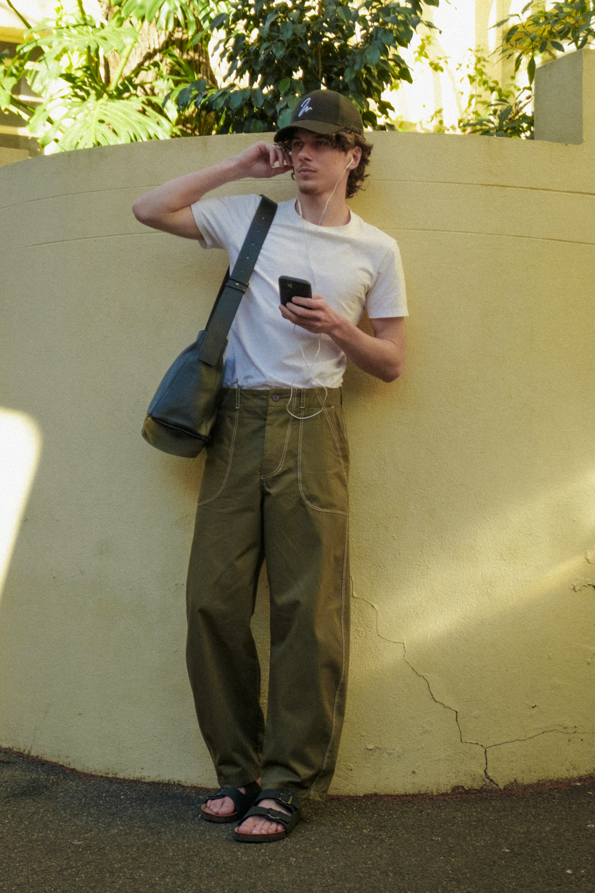 A man stands against a beige stucco wall outdoors, wearing a white t-shirt, olive green fatigue trousers, a black trucker cap, and sandals. He has a black shoulder bag slung over his shoulder and is holding a black smartphone.