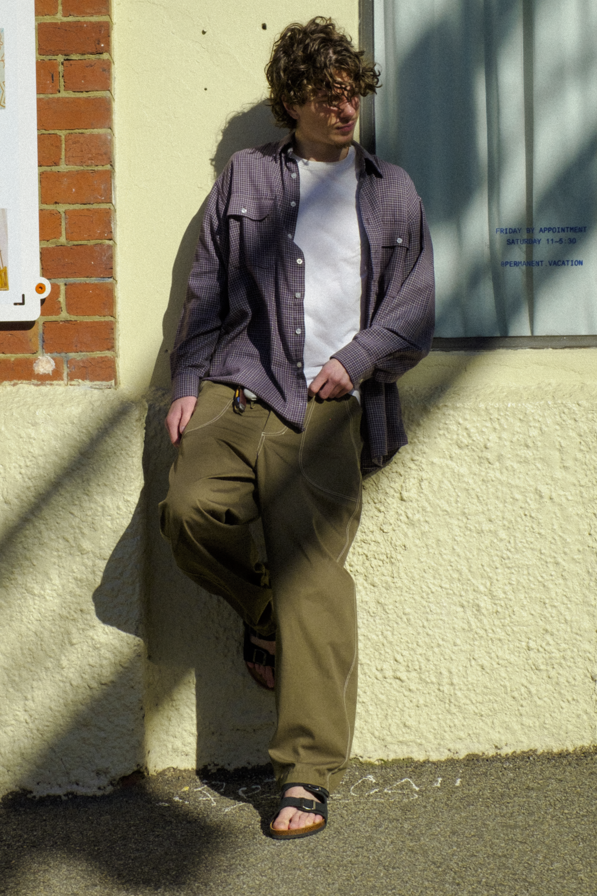 A man wearing a button-down shirt over a white t-shirt, olive green Fatigue Trousers, and sandals, leaning against a textured cream wall outdoors.