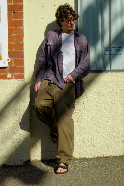 A man wearing a button-down shirt over a white t-shirt, olive green Fatigue Trousers, and sandals, leaning against a textured cream wall outdoors.