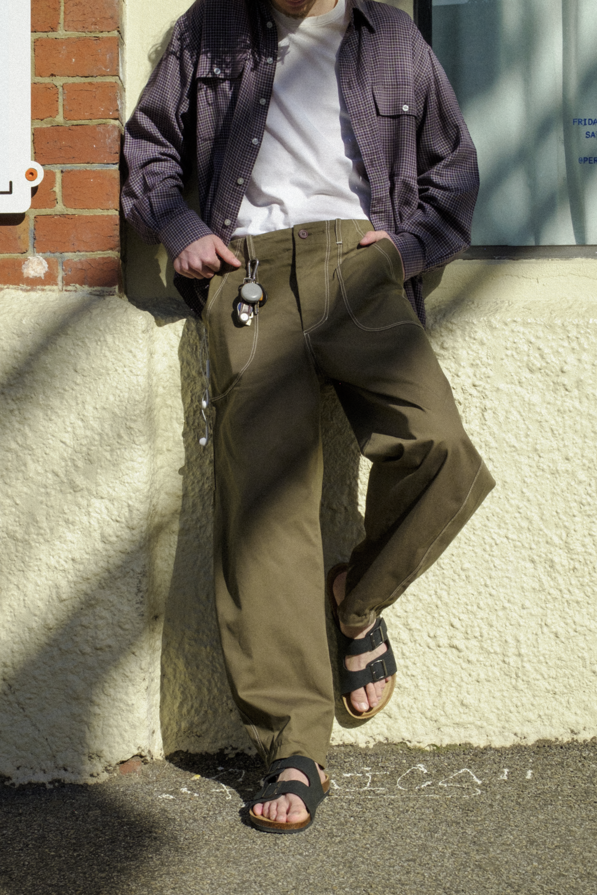A man wearing Fatigue Trousers, a white t-shirt, a  overshirt, and sandals, leaning against a textured cream wall outdoors.