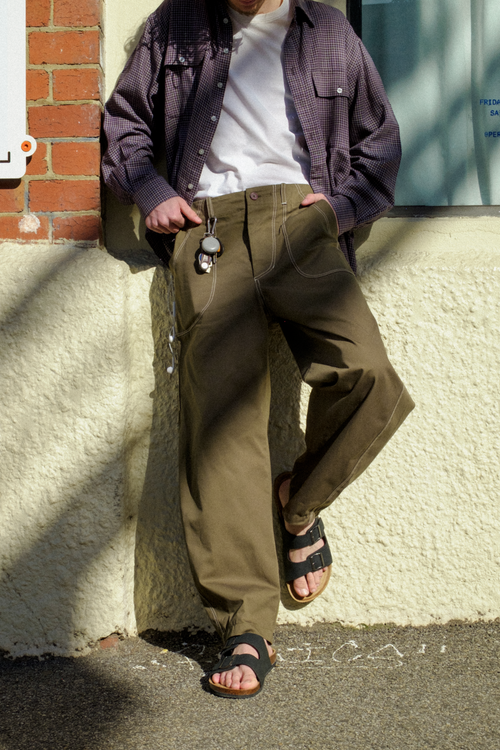 A man wearing Fatigue Trousers, a white t-shirt, a  overshirt, and sandals, leaning against a textured cream wall outdoors.