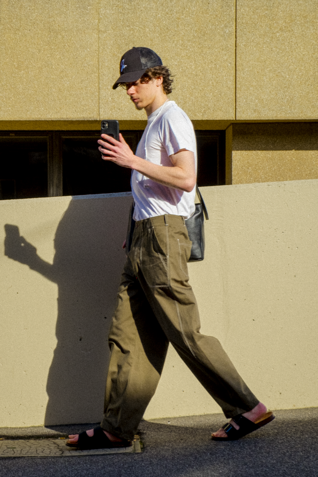 A man walking outdoors while looking at his phone, wearing a white t-shirt, olive fatigue trousers, a black baseball cap, and sandals, with a black shoulder bag.