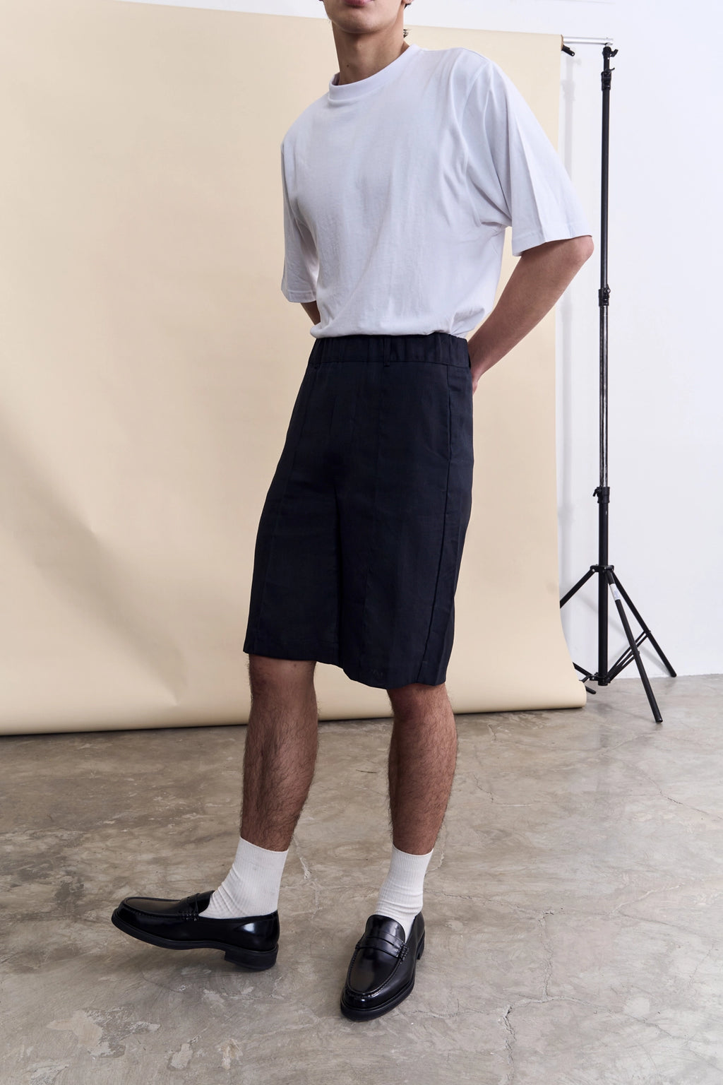 A model wearing a white t-shirt, black knee-length shorts, white socks, and black leather loafers, standing in a studio with a beige backdrop.