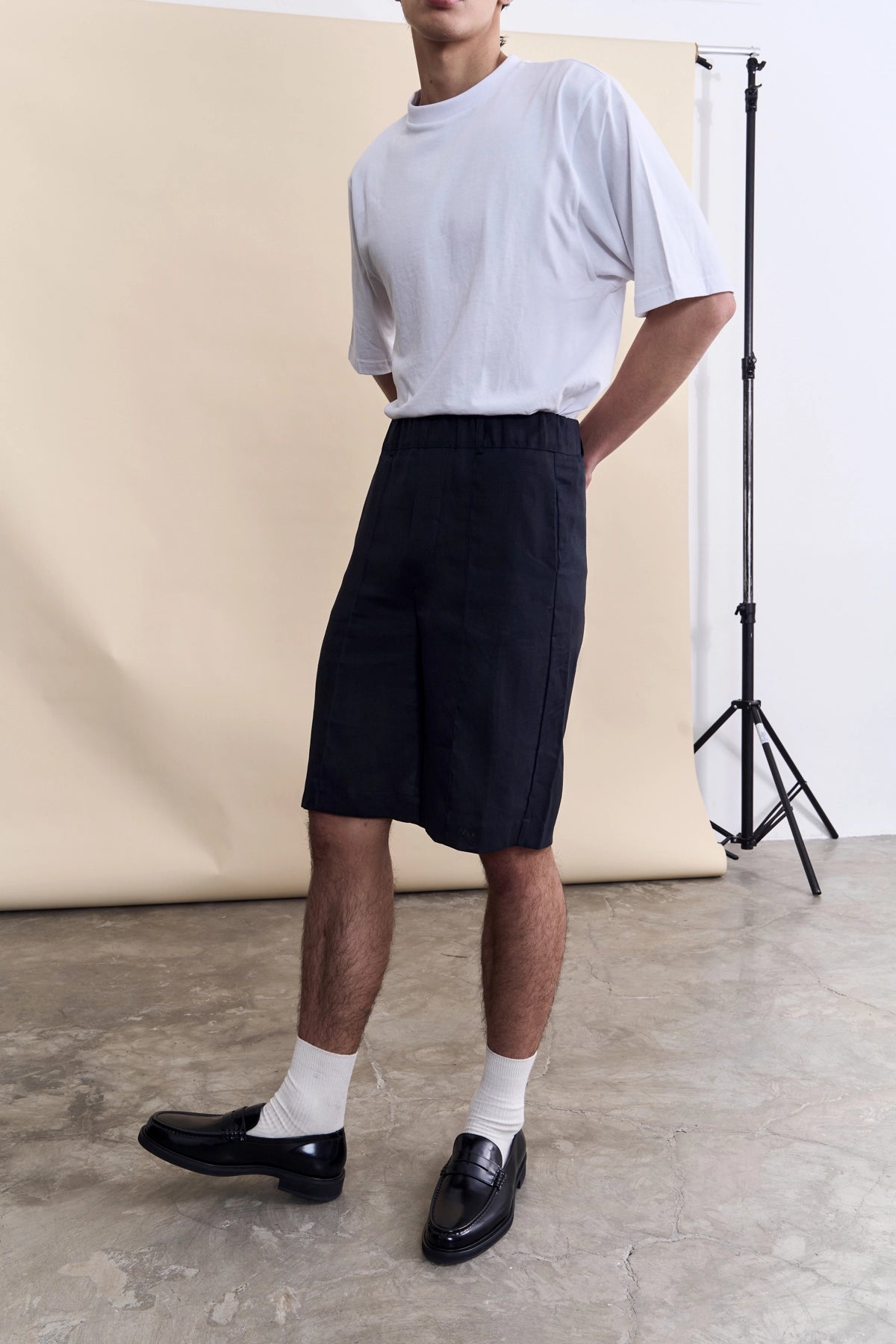 A model wearing a white t-shirt, black knee-length shorts, white socks, and black leather loafers, standing in a studio with a beige backdrop.