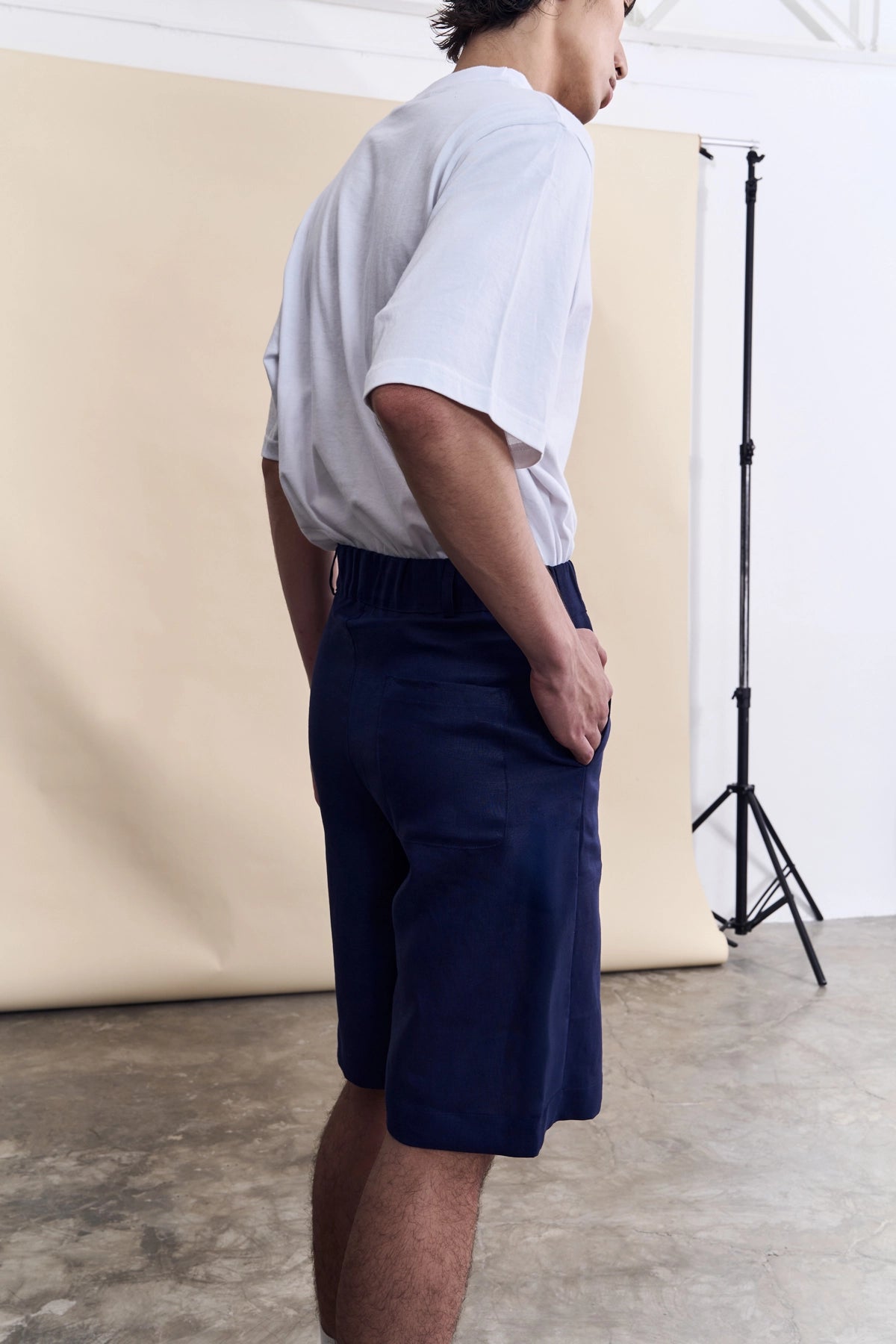 A man wearing navy blue shorts and a white t-shirt, standing in profile in a studio setting with a beige backdrop.