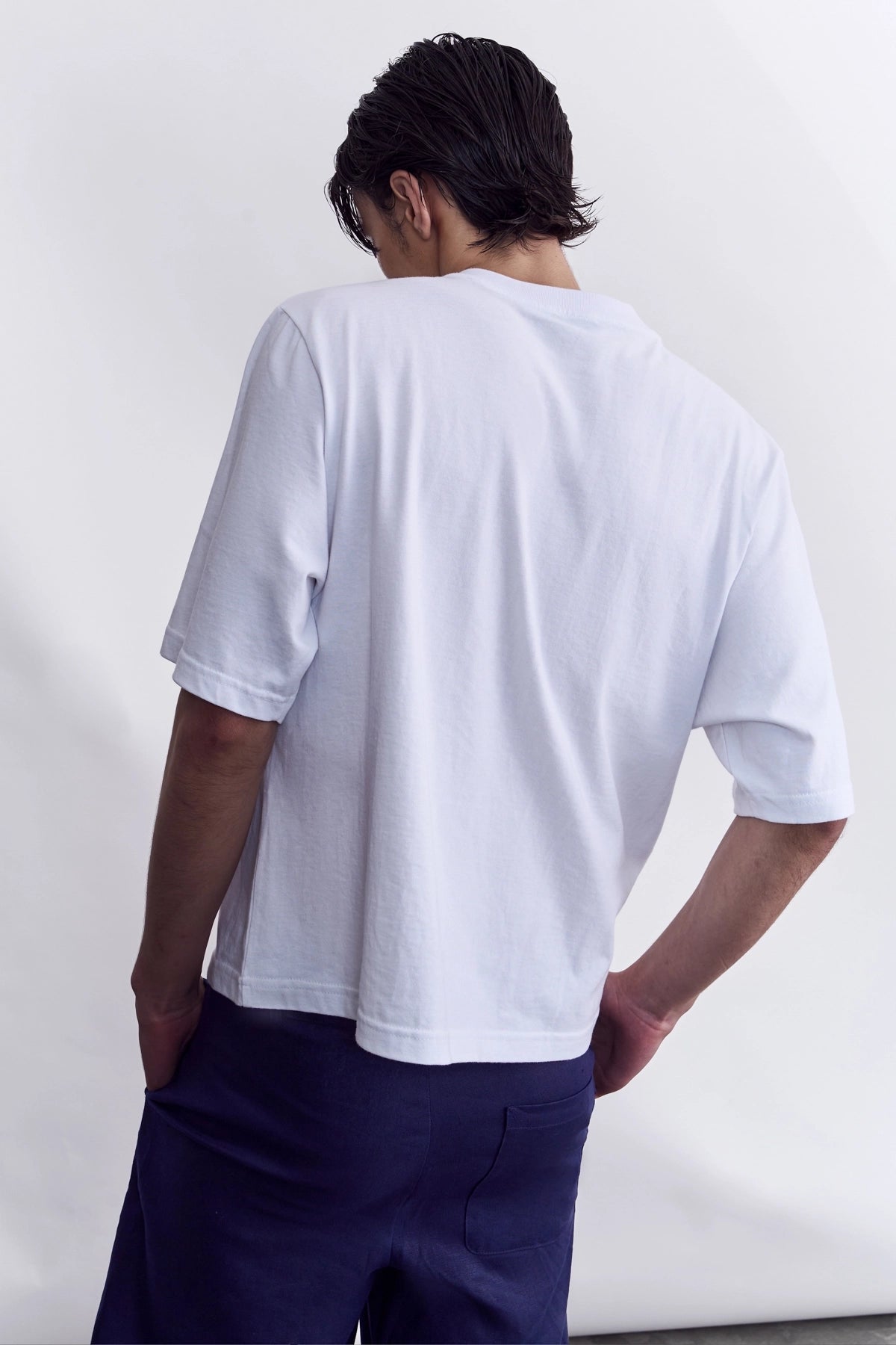A man shown from behind wearing a white short-sleeve t-shirt and navy blue shorts, standing against a plain white studio background.